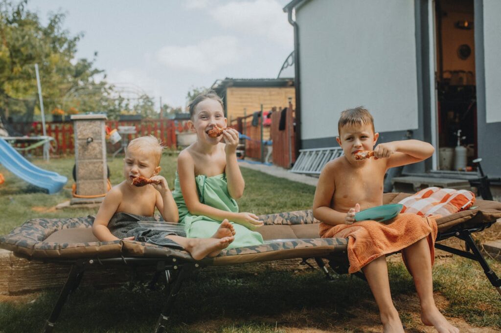 drei Kinder essen genüsslich draußen auf der Liege in Badesachen