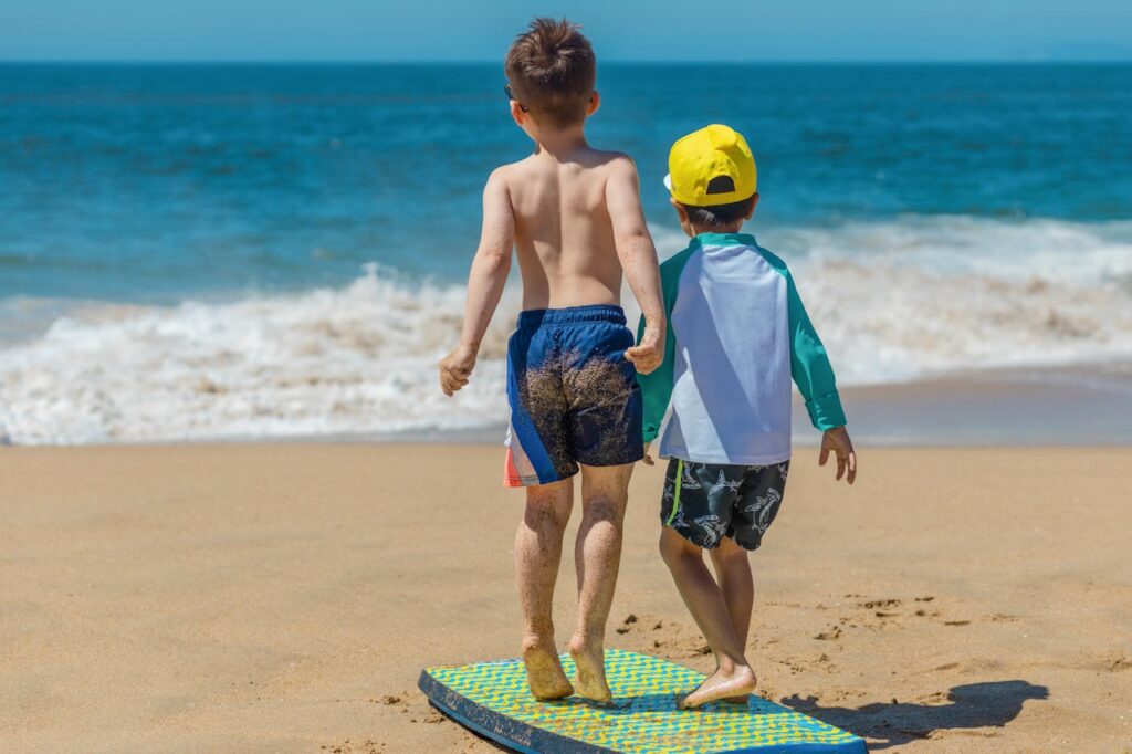 zwei kleine Jungs in Badekleidung stehen auf einem Bodyboard am Strand und schauen auf das Meer
