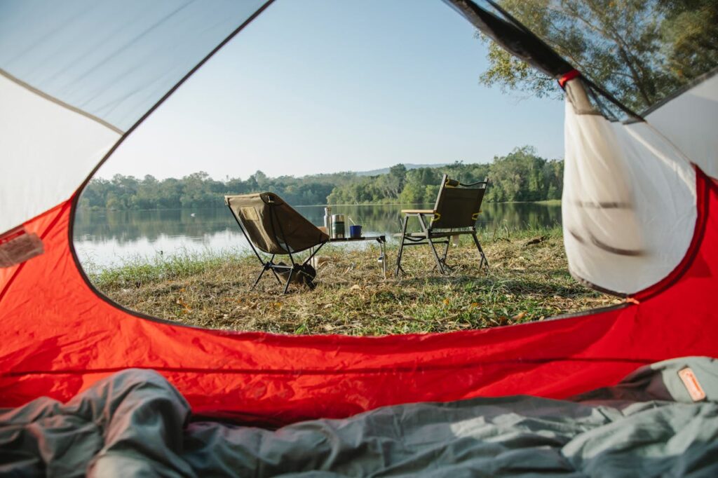 Blick von innen aus einem Zelt auf zwei Campingstühle mit Campingtisch und Wasserflasche an einem See
